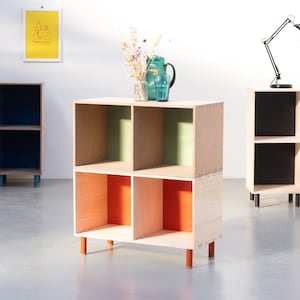 May include: Four wooden cube shelves with different colored interiors. The shelves are stacked on top of each other and have wooden legs. The shelves are painted yellow, blue, green, and orange.
