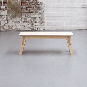 Puede incluir: Un banco de madera clara con un dise&ntilde;o sencillo. El banco tiene una tapa rectangular y cuatro patas. Las patas est&aacute;n inclinadas hacia adentro y tienen una ligera curva en la parte inferior.