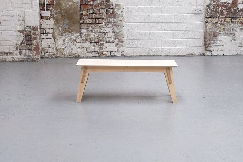 Puede incluir: Un banco de madera clara con un dise&ntilde;o sencillo. El banco tiene una tapa rectangular y cuatro patas. Las patas est&aacute;n inclinadas hacia adentro y tienen una ligera curva en la parte inferior.