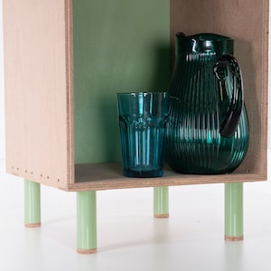 May include: A small wooden shelf unit with green painted legs and a green back panel. The shelf is holding a blue glass and a green glass pitcher.