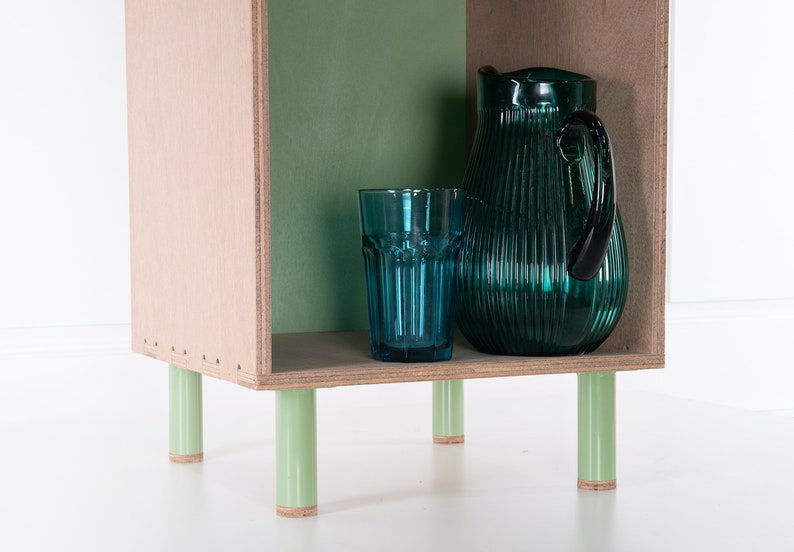 May include: A small wooden shelf unit with green painted legs and a green back panel. The shelf is holding a blue glass and a green glass pitcher.