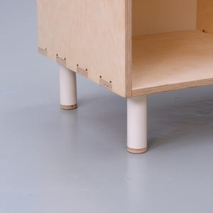 May include: A wooden shelf unit with four white legs. The shelf is made of plywood and has a simple, modern design.