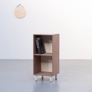 May include: A two-shelf wooden bookcase with white legs. The bookcase is brown and has a light brown interior. There are three books on the top shelf.