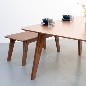 Puede incluir: Un conjunto de mesa y banco de madera con un acabado marr&oacute;n claro. La mesa tiene una tapa cuadrada y cuatro patas. El banco tiene una tapa rectangular y cuatro patas. La mesa y el banco est&aacute;n hechos de madera contrachapada.