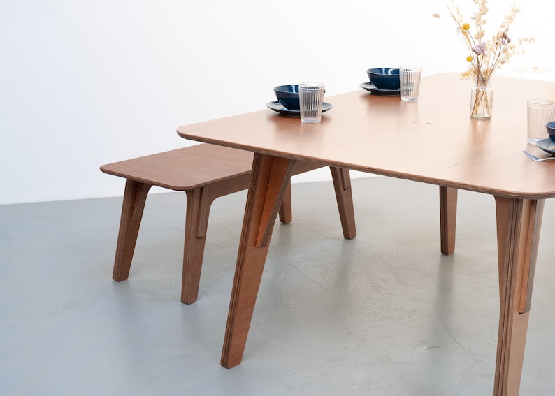 Puede incluir: Un conjunto de mesa y banco de madera con un acabado marr&oacute;n claro. La mesa tiene una tapa cuadrada y cuatro patas. El banco tiene una tapa rectangular y cuatro patas. La mesa y el banco est&aacute;n hechos de madera contrachapada.