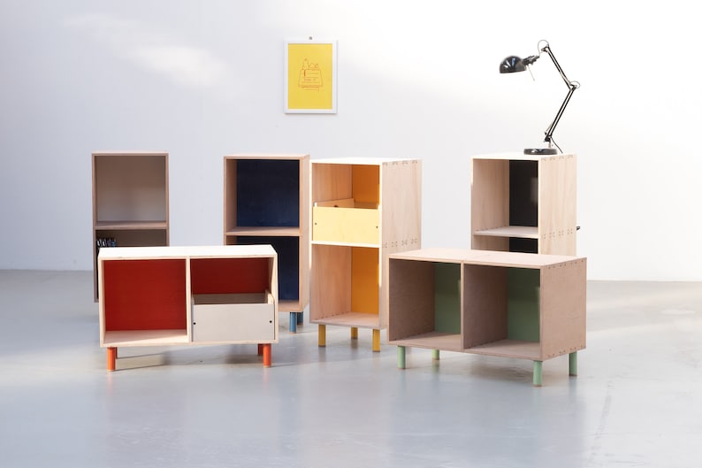 May include: Five wooden storage shelves with different colored interiors. The shelves are on legs and have two shelves each. The shelves are in a white room with a black lamp on a shelf in the background.