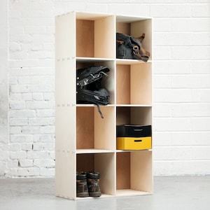 May include: A white wooden shelf unit with nine square compartments. The shelf unit is empty except for a black motorcycle helmet in one compartment, a yellow and black box in another compartment, and a brown leather bag in the top compartment.