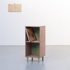 May include: A two-shelf wooden storage unit with a light brown finish and green accents. The unit has four legs and is holding a stack of vinyl records.