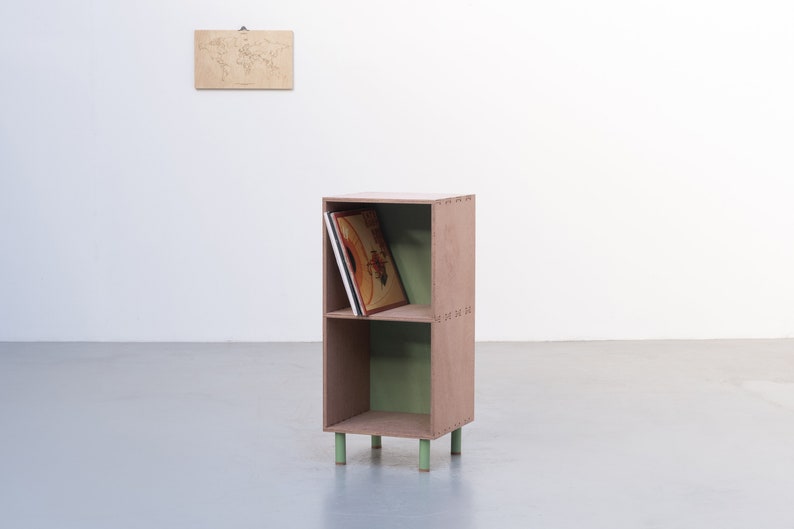 May include: A two-shelf wooden storage unit with a light brown finish and green accents. The unit has four legs and is holding a stack of vinyl records.