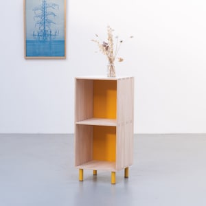 May include: A small wooden shelf unit with two shelves and yellow painted back panels. The shelf unit has four yellow legs and is standing on a gray floor.