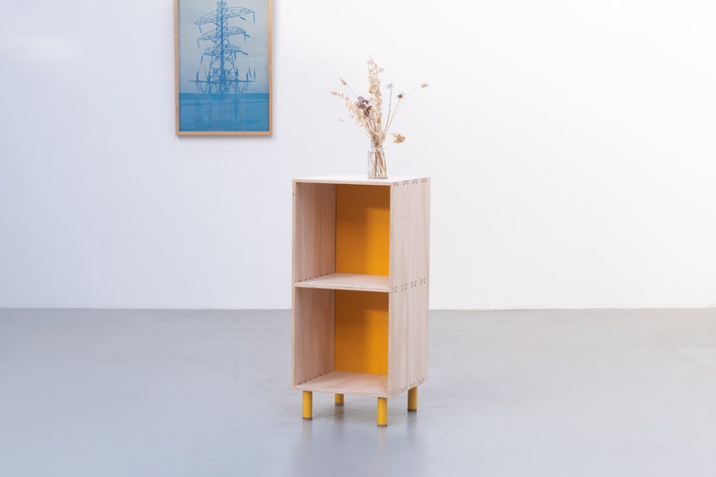 May include: A small wooden shelf unit with two shelves and yellow painted back panels. The shelf unit has four yellow legs and is standing on a gray floor.