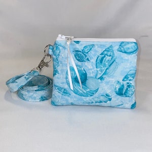 May include: A light blue zippered pouch featuring a repeating pattern of various seashells in darker blue tones. The pouch includes a matching fabric wrist strap attached with a silver-toned metal swivel clasp and a white zipper with a white ribbon pull.