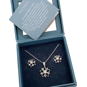 Könnte beinhalten: Ein silberfarbenes Schmuckset mit Halskette und passenden Ohrringen, jeweils mit floralem Design und funkelnden Akzenten. Das Set wird in einer blauen Schachtel mit einer Karte mit der Aufschrift "PAST TIMES Victorian (1837-1901)" präsentiert.