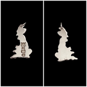 May include: A silver pendant in the shape of the UK, with a small loop for a chain. The pendant has hallmarks at the bottom. The pendant is displayed against a black background.