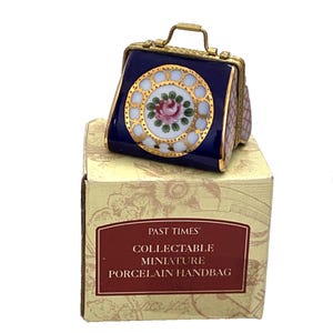 Pode incluir: Uma bolsa em miniatura de porcelana com acabamento azul escuro, apresentando um medalhão circular branco central com uma rosa rosa e folhas verdes, bordas douradas e uma alça de metal, sobre uma caixa com a etiqueta Past Times Collectable Miniature Porcelain Handbag.