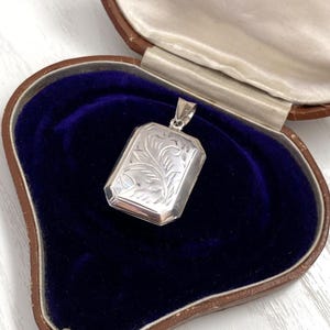 Op de afbeelding: Een rechthoekige zilveren hanger met een gegraveerd bloemmotief, gepresenteerd in een vintage bruine sieradendoos met een blauw fluwelen interieur. De hanger heeft een kleine lus voor een ketting.
