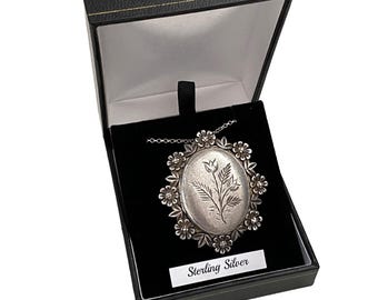 Puede incluir: Colgante de plata con diseño floral, presentado en una caja de joyería negra. El colgante presenta un centro ovalado con un grabado floral, rodeado por un borde floral. La caja incluye una etiqueta que dice "Sterling Silver".