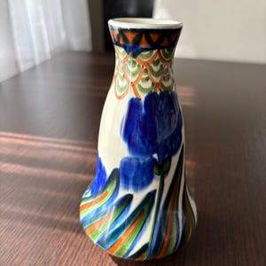 May include: A ceramic vase with a cream-coloured base featuring hand-painted blue iris flowers, green leaves, and orange accents. The neck of the vase is decorated with a geometric pattern of orange triangles and green scalloped lines.