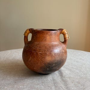 May include: A round, brown, mottled gourd vessel featuring two handles wrapped in light-coloured, thin, coiled material. The surface of the gourd is decorated with fine, etched, white line drawings of floral patterns and dark, smoky shading.