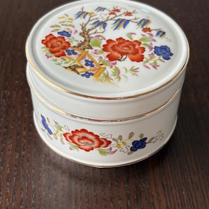 May include: A round white porcelain trinket box with a lid featuring a colorful floral pattern of red and blue flowers with green leaves. The box has gold trim along the edges of the lid and the base, and a matching floral design around the side.