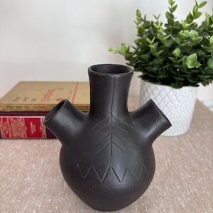 Peut inclure: Un vase en céramique noir mat doté d'un col central et de deux becs latéraux plus petits. La surface présente des motifs décoratifs gravés, notamment un dessin en forme de feuille sur le col et une ligne en zigzag entourant la base arrondie.