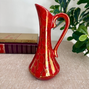 May include: A glossy red ceramic pitcher featuring a repeating pattern of circular shapes in a lighter shade of red. The pitcher has a tall, slender neck, a wide base, and a curved handle extending from the upper neck to the body of the vessel.