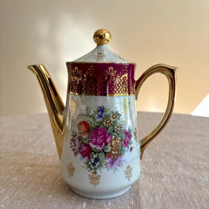 May include: A porcelain teapot featuring a white base with a vibrant floral bouquet of pink, red, and purple roses. The upper rim is a deep burgundy band with intricate gold filigree. It includes a gold-toned handle, spout, and lid finial.