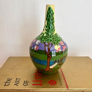 May include: A ceramic vase with a narrow neck and a bulbous body, featuring a hand-painted landscape scene with a green tree, a blue river, a small wooden bridge, and colourful lanterns against a purple sky, all set on a cream-coloured background.