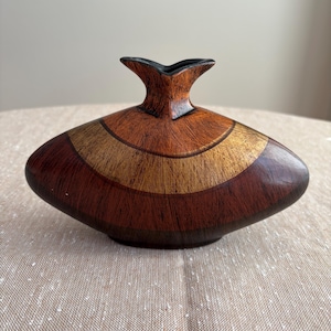 May include: A low-profile, wide-bodied vase with a flattened, elliptical shape. The surface features a wood-grain texture with concentric bands of dark brown, medium brown, and a lighter golden-tan colour. It has a small, flared, black-rimmed opening.