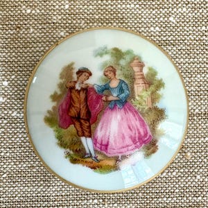 May include: A round porcelain trinket box lid featuring a colourful illustration of two figures in period clothing dancing in a garden setting. The scene includes a person in a brown coat and pink cape and another in a blue jacket and pink skirt, with the name Fragonard printed near the bottom edge, all framed by a thin gold metal rim.