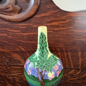 May include: A small, bulbous ceramic vase with a narrow neck, featuring a hand-painted landscape scene. The design includes a prominent green tree with textured leaves, a purple sky, a blue river, and pink floral accents against a yellow background.