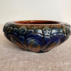 May include: A low, round ceramic bowl featuring a deep blue and brown reactive glaze. The exterior surface is decorated with a repeating embossed floral or petal pattern that wraps around the entire circumference of the vessel.