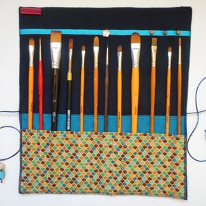 Peut inclure: Un étui à pinceaux en tissu bleu marine avec un motif géométrique coloré en bas. L'étui contient divers pinceaux avec des manches orange et des viroles argentées. Un cordon bleu avec une perle en bois et une perle turquoise est attaché.
