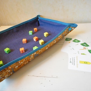 May include: A blue fabric tray with a floral pattern and a blue trim. The tray is filled with colorful dice. The dice are in various colors, including green, orange, and yellow.