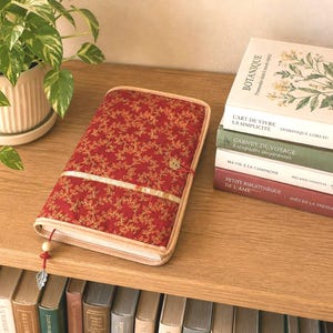 Peut inclure: Une couverture de livre en tissu rectangulaire rouge profond avec un motif de feuilles dorées, dotée d'une bordure beige clair, d'un ruban doré horizontal et d'un marque-page en cordon rouge avec une perle en bois et une breloque feuille en métal argenté.