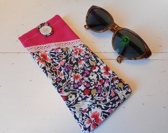 May include: A floral patterned fabric sunglasses case with a pink flap and a white button closure. The case is lying on a white surface next to a pair of brown tortoise shell sunglasses.
