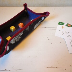 Op de afbeelding: Een donkerblauwe stoffen dobbelsteenbak met een rode rand en een wit stippenpatroon. De bak is gevuld met vier dobbelstenen, twee oranje en twee rood. Aan de rechterkant van de afbeelding zijn drie kaarten te zien, met de tekst "Citron vert confit" en "Jouret de vert".