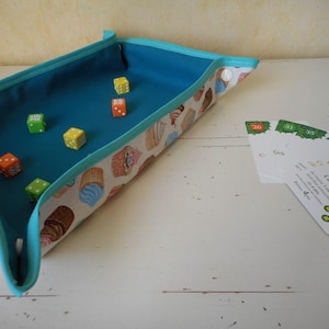 May include: A blue fabric tray with a white and blue trim, filled with colorful dice. The tray has a pattern of cupcakes on it. There are also four cards with the text "Gâteau aux fruits" on them.