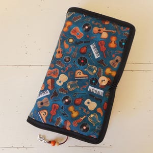 Può includere: Un astuccio rettangolare in tessuto con bordo nero e chiusura a bottone. L'astuccio è decorato con vari strumenti musicali, tra cui chitarre, violini e pianoforti, su uno sfondo verde acqua. Un ciondolo a forma di nota musicale e perline pendono dall'astuccio.