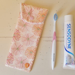 Peut inclure: Un porte-brosse à dents en tissu floral rose et blanc avec une fermeture à bouton rose. Une brosse à dents blanche avec un manche rose et des poils bleus est à côté d'un tube de dentifrice blanchissant Sensodyne.