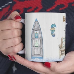 May include: White ceramic mug featuring a nautical theme. The design includes a blue boat, seahorse, and other coastal elements. The mug is held by a person wearing a navy blue sweater with a red and white pattern.
