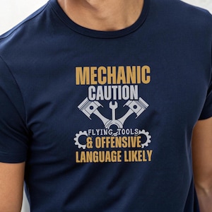 May include: Navy blue t-shirt with a mechanic-themed graphic. The design includes the text "MECHANIC CAUTION" above an illustration of pistons and a wrench. Below, it reads "FLYING TOOLS & OFFENSIVE LANGUAGE LIKELY."