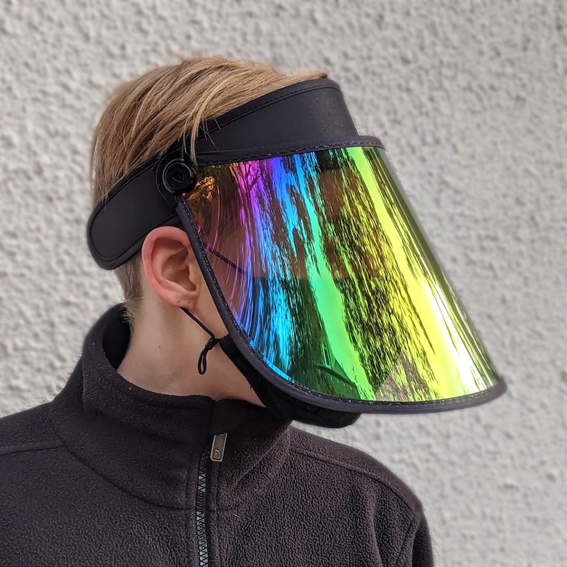 May include: Black visor with a rainbow-coloured, iridescent shield. The visor has a black band and a button detail on the side. A black string is attached to the visor. The visor is worn by a person.