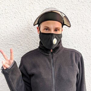 May include: A black face mask with a white and yellow circular design, worn with a black protective visor and a black fleece jacket. The visor has a clear plastic shield. The person is giving a peace sign.