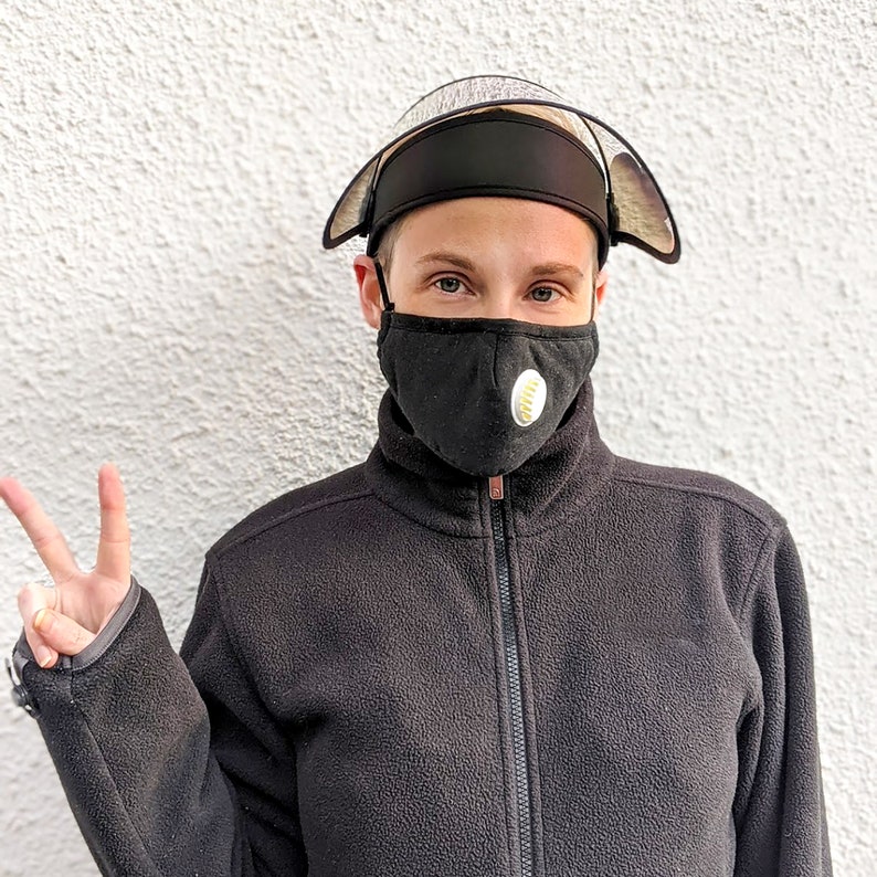 May include: A black face mask with a white and yellow circular design, worn with a black protective visor and a black fleece jacket. The visor has a clear plastic shield. The person is giving a peace sign.