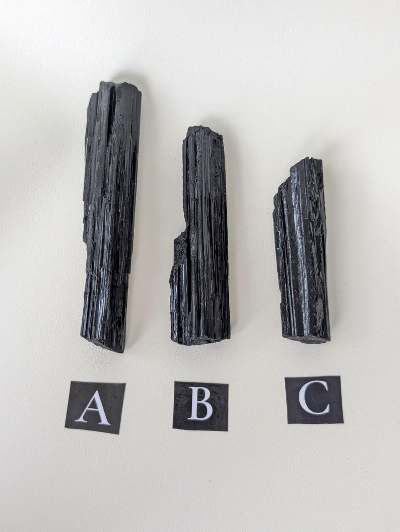 May include: Three black tourmaline crystals of varying sizes are displayed against a white background. Each crystal has a rough, textured surface with vertical striations. Small black cards labeled A, B, and C are placed below the crystals.