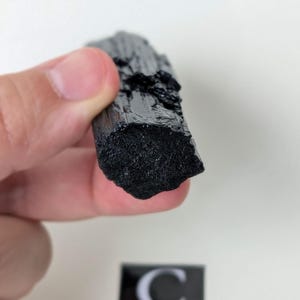 May include: A raw, black tourmaline crystal held in a hand. The crystal has a rough, textured surface and a dark, opaque color. The image includes a small, black and white label with the letter "C".