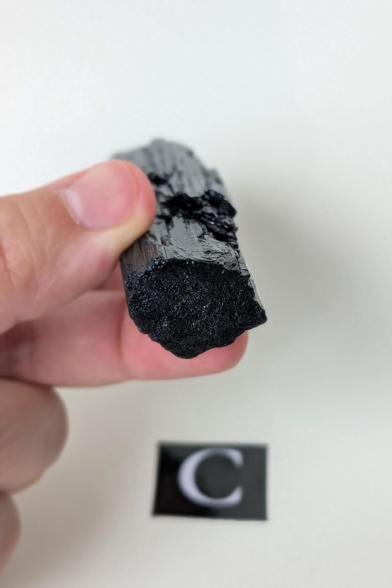 May include: A raw, black tourmaline crystal held in a hand. The crystal has a rough, textured surface and a dark, opaque color. The image includes a small, black and white label with the letter "C".