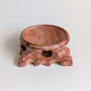 May include: A circular wooden display stand with a reddish-brown grain pattern. The stand features a smooth, recessed top surface for holding objects and a carved, organic base with small cutouts, showcasing a natural wood texture throughout.