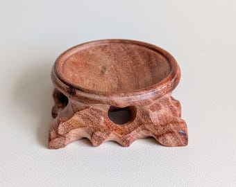 May include: A circular wooden display stand with a reddish-brown grain pattern. The stand features a smooth, recessed top surface for holding objects and a carved, organic base with small cutouts, showcasing a natural wood texture throughout.